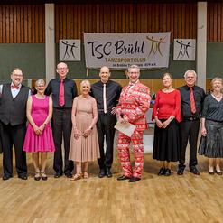 Gruppe von zw&ouml;lf T&auml;nzern in festlicher Kleidung, posierend vor einem Banner des TSC Br&uuml;hl in einer Tanzhalle.