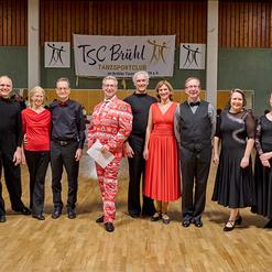 Gruppe von T&auml;nzern in eleganter Kleidung, die in einem Tanzsaal posiert, mit Banner des TSC Br&uuml;hl im Hintergrund.