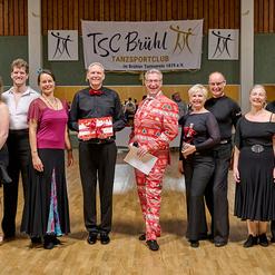 Gruppe von T&auml;nzern in festlicher Kleidung posiert mit Pokalen vor einem Banner des TSC Br&uuml;hl in einer Tanzhalle.