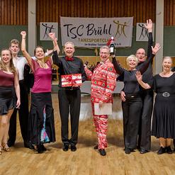 Gruppenszene von tanzenden Personen im Festkleid auf einer Tanzveranstaltung im TSC Br&uuml;hl, alle mit freudigen Gesten.