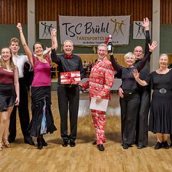 Gruppe von T&auml;nzern in festlicher Kleidung feiert mit einem Pokal in einer Tanzhalle. Banner des TSC Br&uuml;hl im Hintergrund.