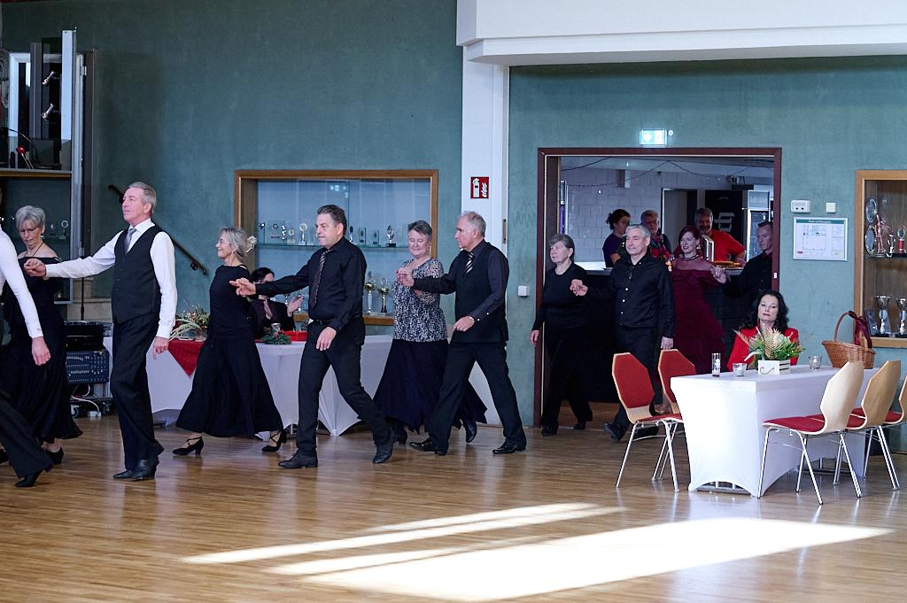 Tanzende Personen in eleganter Kleidung in einem festlichen Saal mit Tischen im Hintergrund.