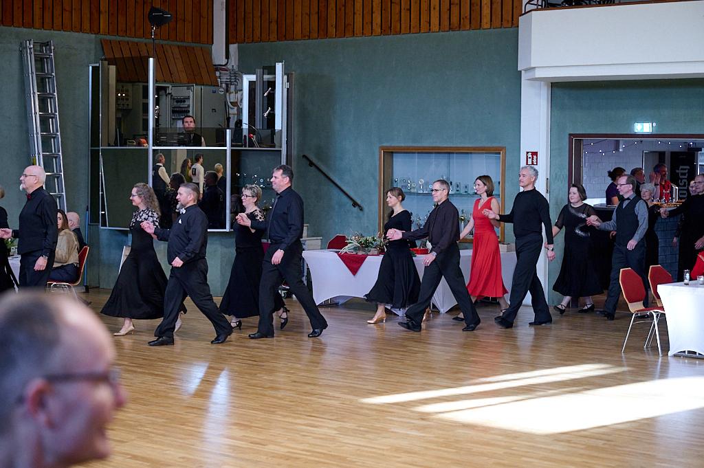Tanzende Paare in eleganter Kleidung gehen in einer Festhalle aufeinander zu. Tische sind im Hintergrund sichtbar.