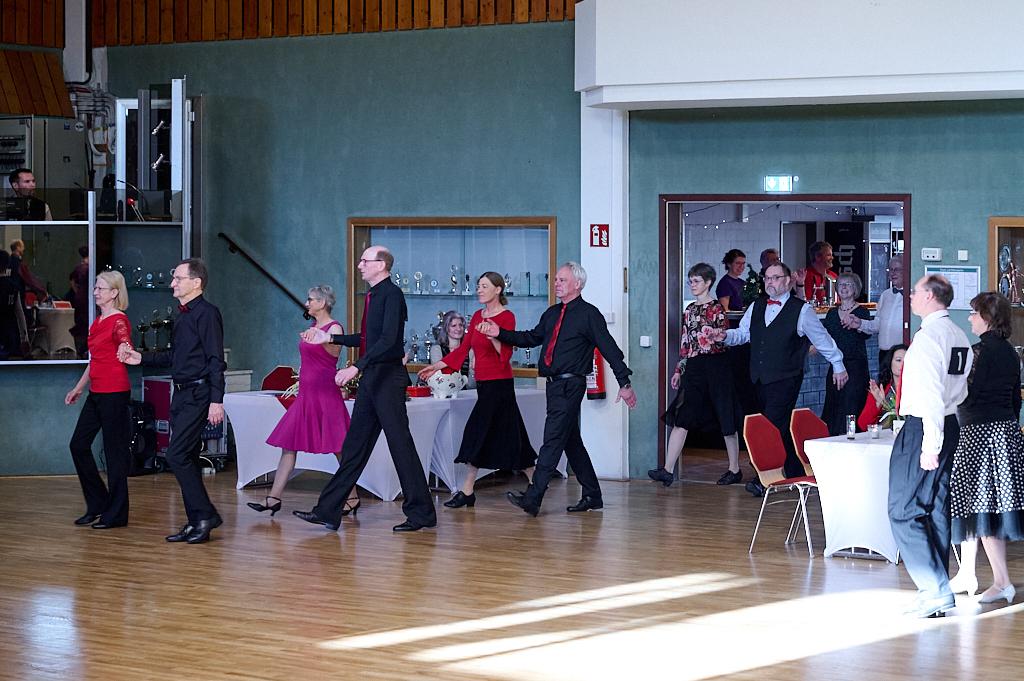 Tanzende Paare in eleganter Kleidung bewegen sich in einem festlichen Saal mit Tischen im Hintergrund.