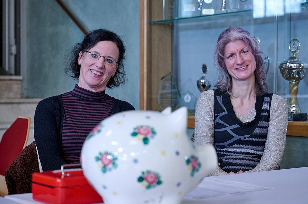 Zwei Frauen l&auml;cheln am Tisch, ein buntes Sparschwein und eine rote Box sind sichtbar im Vordergrund.