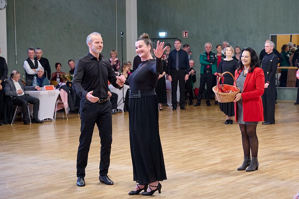 Tanzpaar in festlicher Kleidung, eine Frau winkt, w&auml;hrend eine andere mit einem Korb im Hintergrund l&auml;chelt. Zuschauer beobachten.