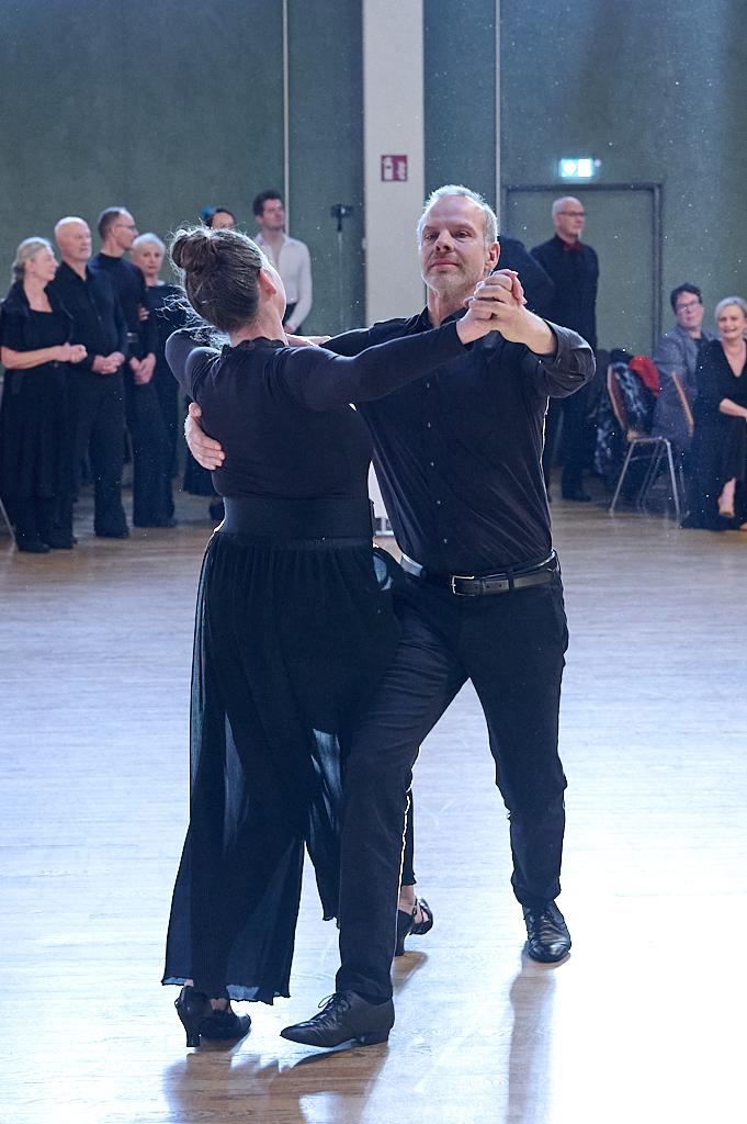 Tanzpaar in eleganter Kleidung f&uuml;hrt einen Tanz auf einer Halle mit Zuschauern im Hintergrund aus.