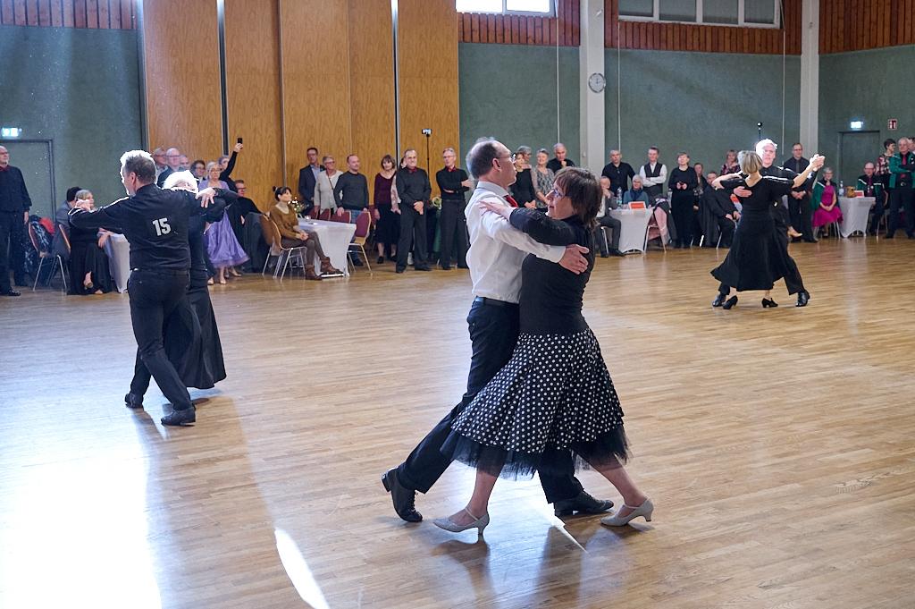 Tanzpaar im Halbprofil auf einer Tanzfl&auml;che, umgeben von Zuschauern in festlicher Kleidung.