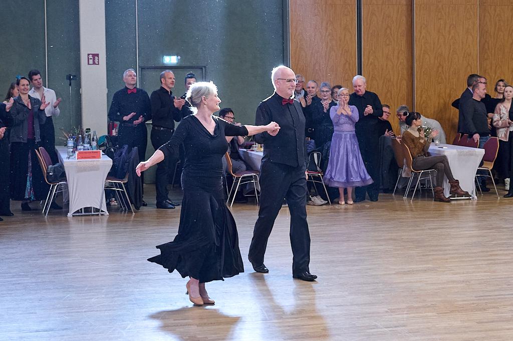 Paar tanzt in festlicher Kleidung auf einer Tanzfl&auml;che, umgeben von applaudierenden Zuschauern in einem Veranstaltungssaal.
