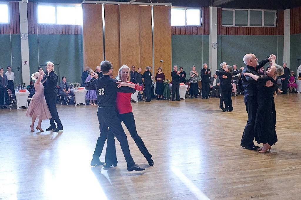 Tanzpaar in eleganter Kleidung tanzt auf einer Holzfl&auml;che, umgeben von Zuschauern in einem festlichen Saal.