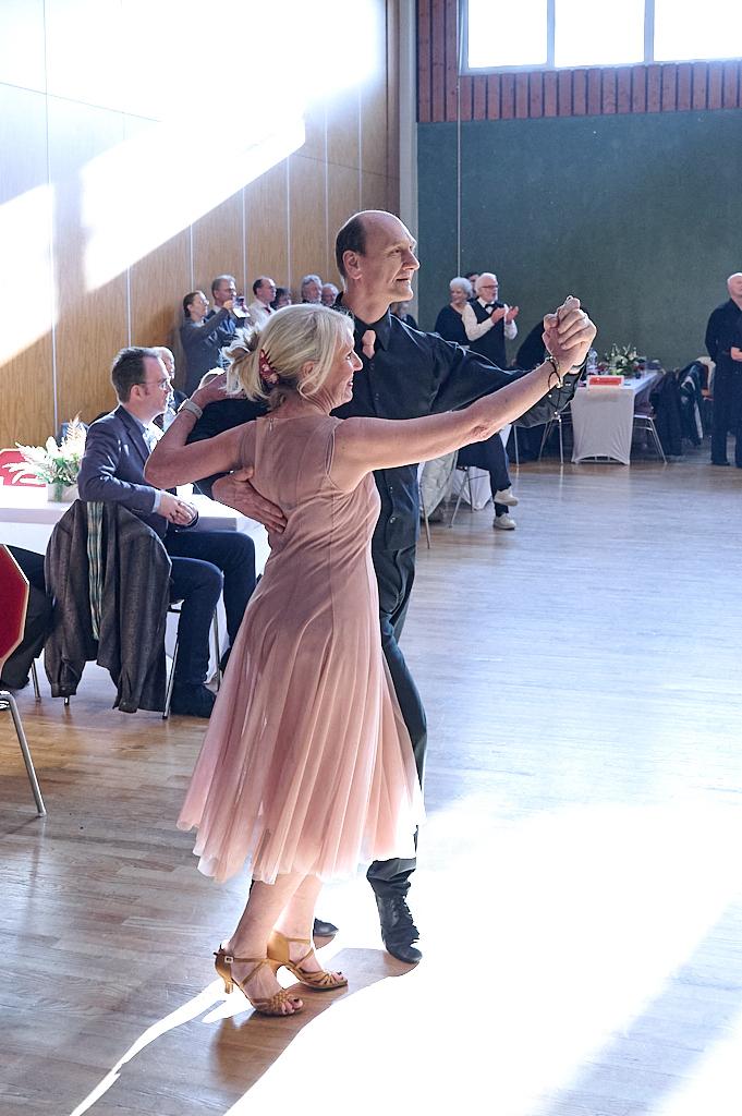 Tanzendes Paar auf einer Veranstaltung, umgeben von Publikum, in einem hellen Saal mit Holzvert&auml;felung.