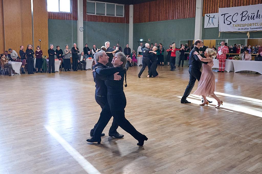 Tanzpaare in eleganter Kleidung beim Tanzen in einem festlichen Saal mit Publikum im Hintergrund.