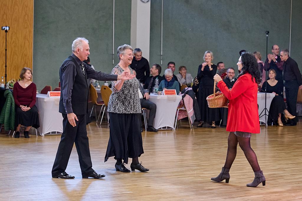 Tanzpaar auf einer Veranstaltung, w&auml;hrend eine Frau in rotem Mantel einen Korb h&auml;lt, Zuschauer im Hintergrund.