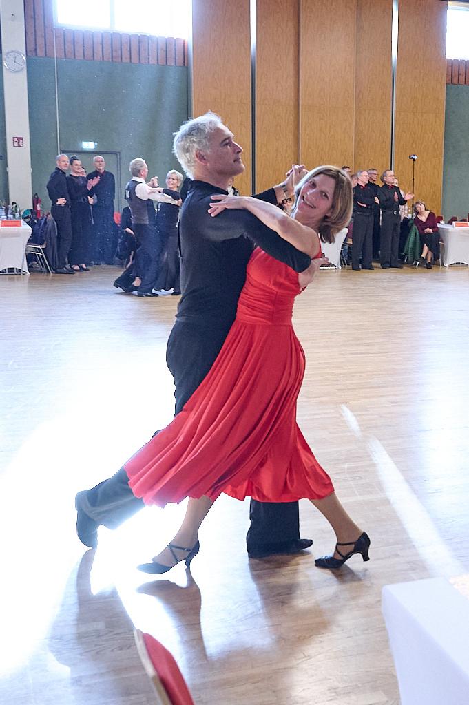 Tanzpaar in eleganter Kleidung f&uuml;hrt einen Tanz in einem festlichen Saal mit anderen T&auml;nzern im Hintergrund aus.