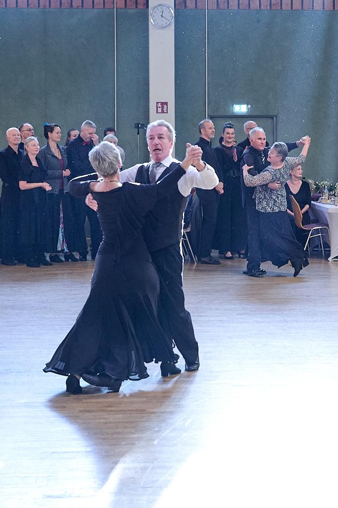 Tanzendes Paar in eleganter Kleidung auf einer Tanzfl&auml;che, umgeben von Zuschauern in festlicher Kleidung.