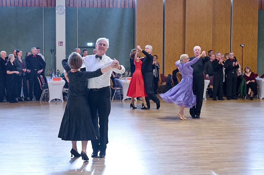 Tanzpaare in eleganter Kleidung tanzen auf einer Tanzfl&auml;che, G&auml;ste beobachten und applaudieren im Hintergrund.