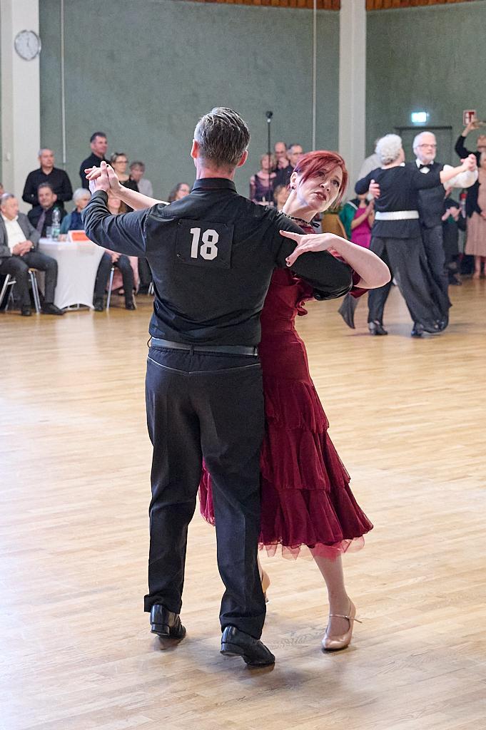 Tanzpaar in festlicher Kleidung f&uuml;hrt einen modernen Tanz in einem Saal mit Zuschauern im Hintergrund aus.