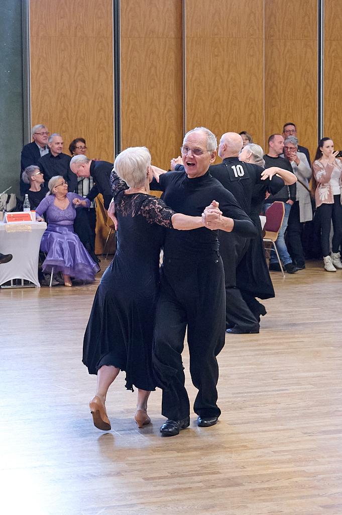 Tanzendes Paar im Schwarzen, umgeben von Zuschauern in einem festlichen Saal. Holzfu&szlig;boden und gro&szlig;e Fenster im Hintergrund.
