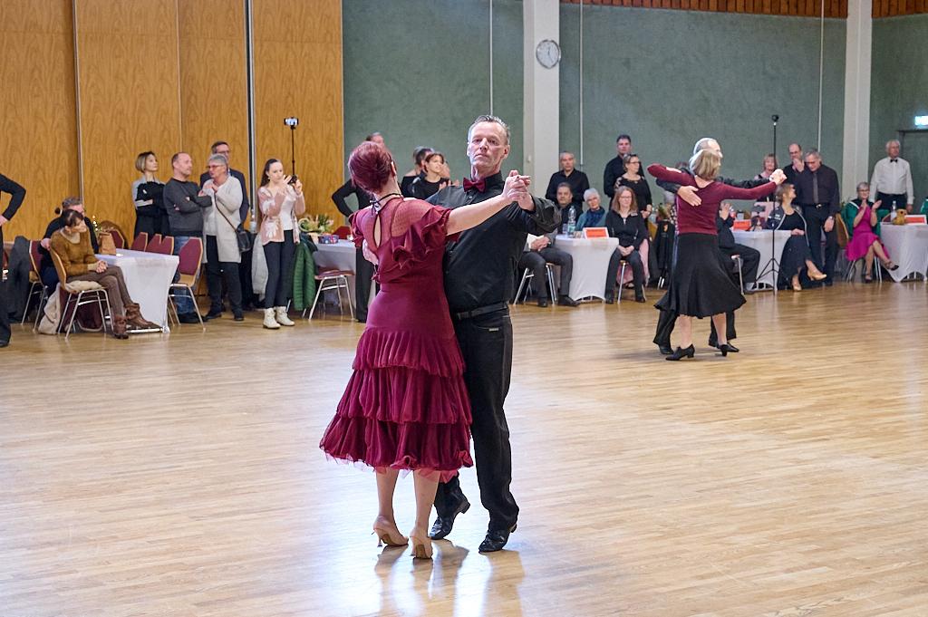 Tanzpaar in eleganter Kleidung tanzt in einem Saal, umgeben von zuschauenden Personen. Holzfu&szlig;boden und Dekor sichtbar.