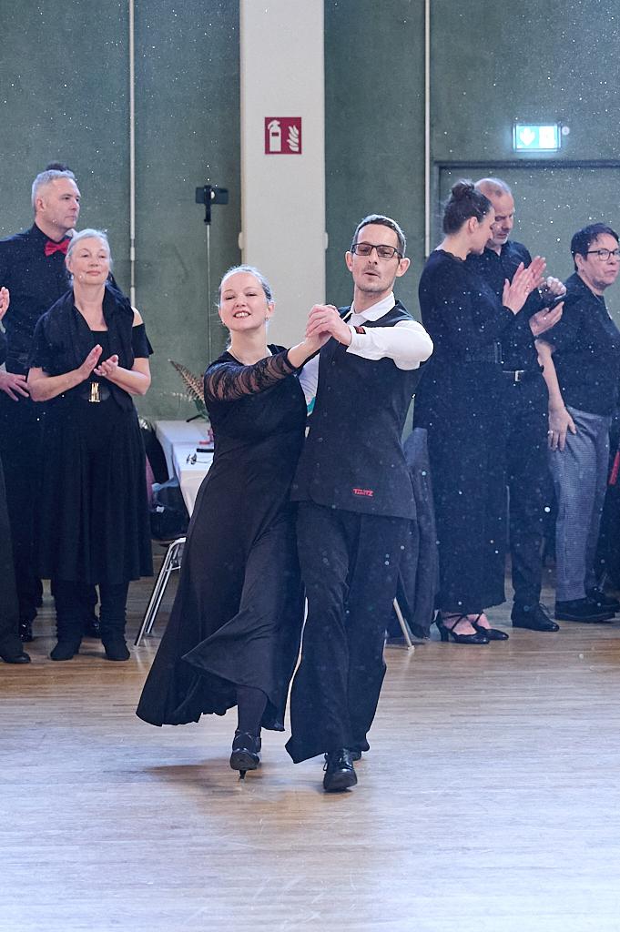 Tanzpaar in eleganter Kleidung f&uuml;hrt einen Tanzschritt vor applaudierendem Publikum in einem festlichen Raum aus.