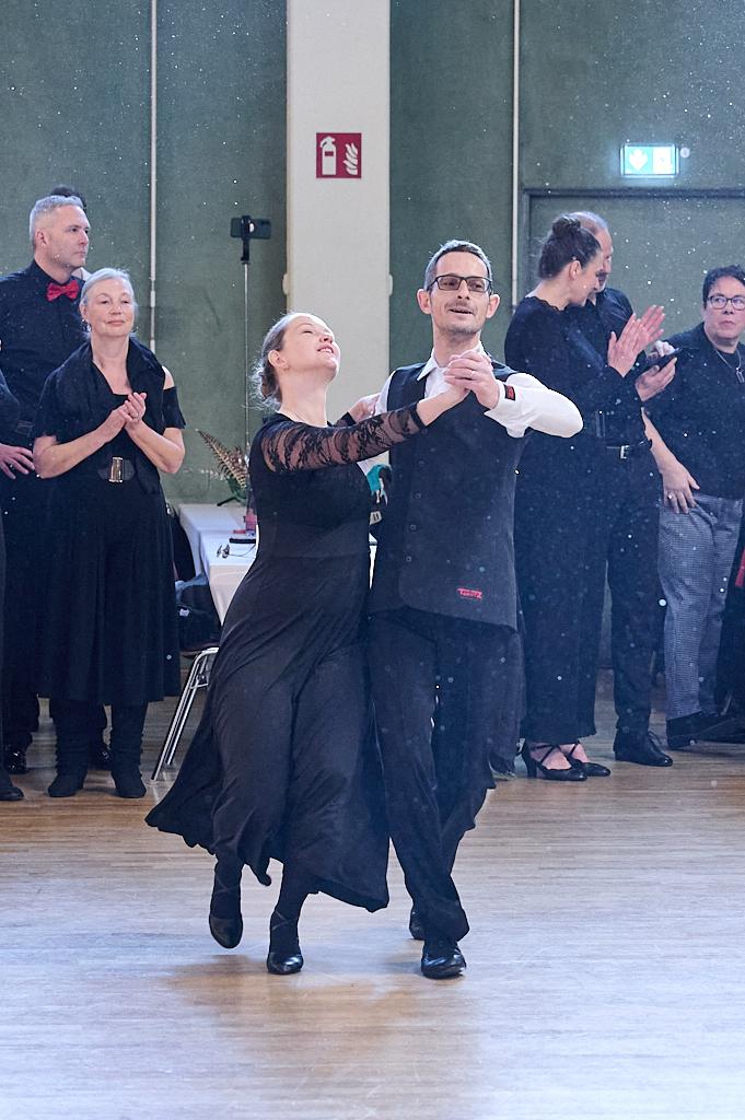 Tanzpaar in eleganter Kleidung f&uuml;hrt einen Schritt auf der Tanzfl&auml;che, umgeben von applaudierenden Zuschauern.