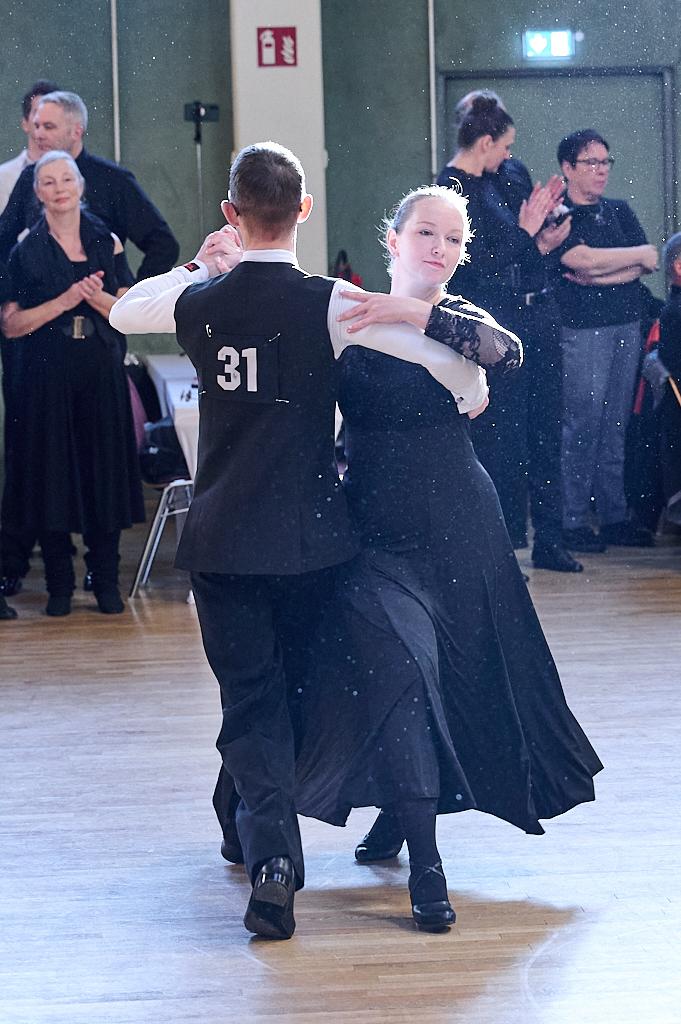 Tanzpaar in eleganter Kleidung tanzt im Fokus auf einer Tanzfl&auml;che, Zuschauer applaudieren im Hintergrund.