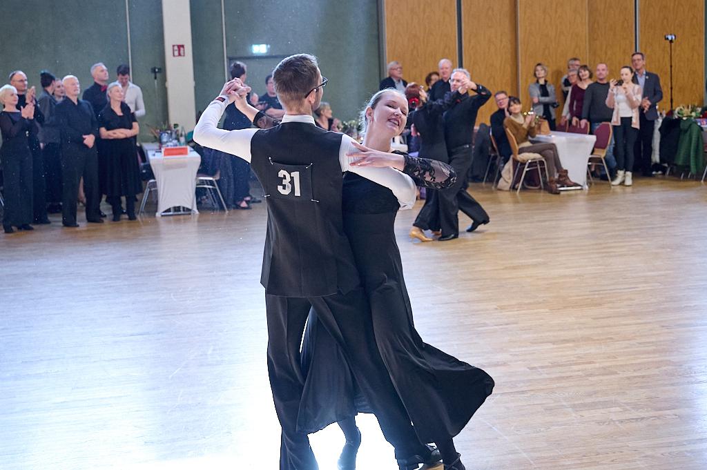 Tanzpaar in festlicher Kleidung tanzt auf einer Tanzfl&auml;che, Zuschauer applaudieren im Hintergrund.