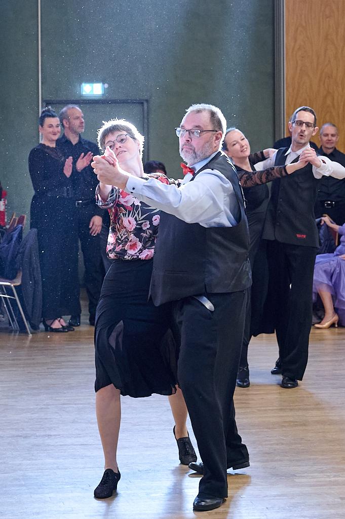 Tanzpaar in eleganter Kleidung f&uuml;hrt einen Walzer auf einer Tanzfl&auml;che, Zuschauer applaudieren im Hintergrund.
