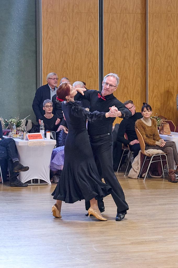 Tanzendes Paar in eleganter Kleidung, umgeben von Zuschauern in einem festlichen Saal mit Holzboden.