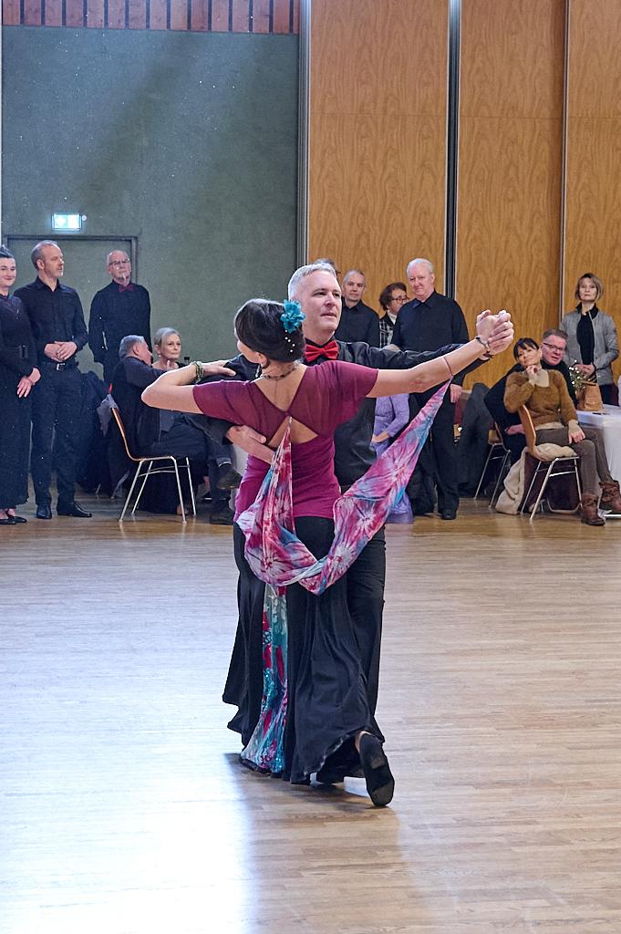 Tanzenden Paar in eleganter Kleidung, umgeben von Zuschauern in einem Tanzsaal mit Holzfu&szlig;boden.