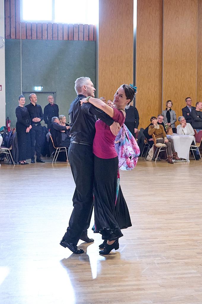 Tanzpaar in eleganter Kleidung tanzt auf einer hellen Tanzfl&auml;che, Zuschauer im Hintergrund beobachten aufmerksam.