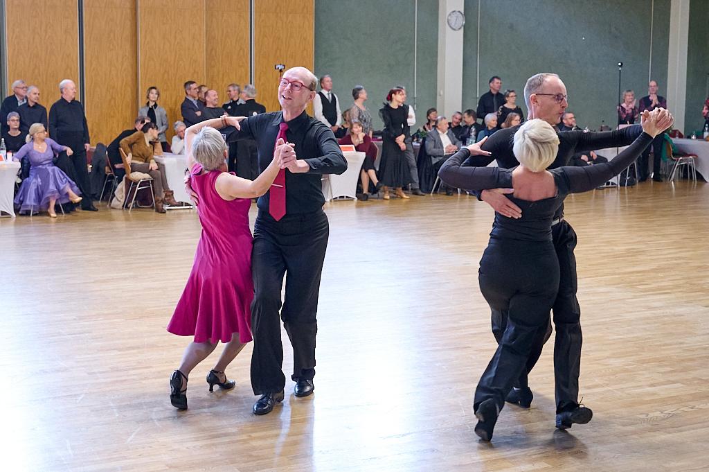 Tanzveranstaltung in einem Saal: Paare tanzen in eleganter Kleidung auf einem Parkettboden, Zuschauer im Hintergrund.