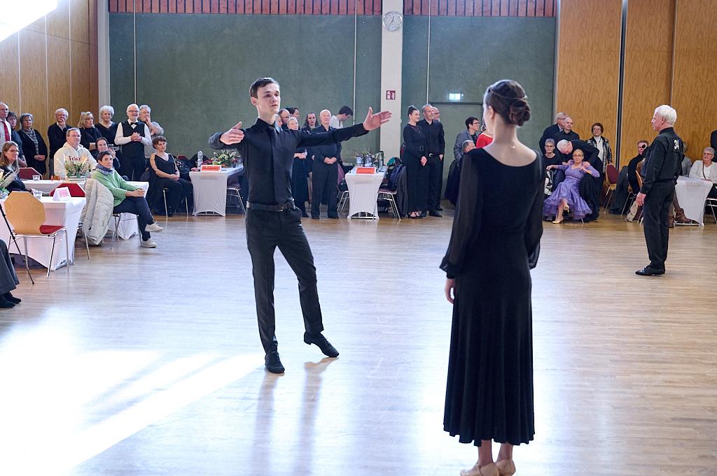 Tanzpaar auf einer Fl&auml;che, Mann in schwarzem Outfit dirigiert Frau in schwarzem Kleid vor Publikum in festlicher Kleidung.