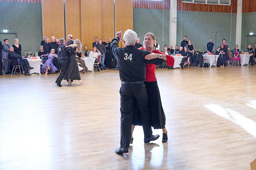 Tanzpaar in eleganter Kleidung tanzt auf einer Holzfl&auml;che, Zuschauer sitzen an Tischen im Hintergrund.