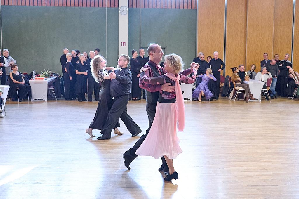 Tanzpaar in eleganter Kleidung tanzt in einem hellen Saal, umgeben von Zuschauern an festlich gedeckten Tischen.