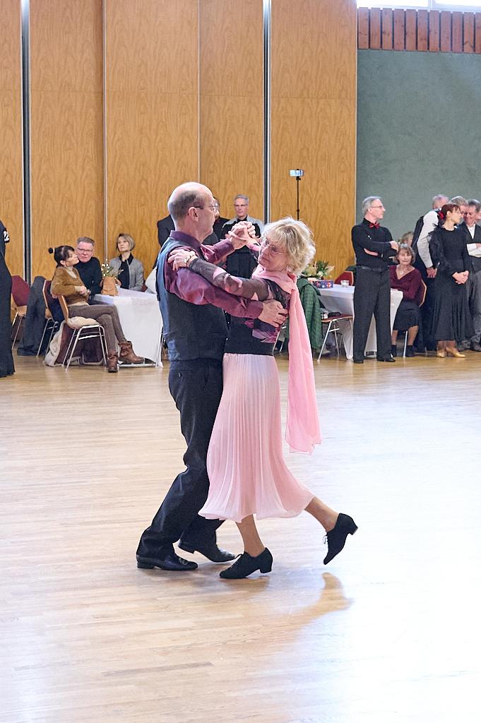 Tanzendes Paar auf einer Veranstaltung, umgeben von Zuschauern in einem gro&szlig;en Saal mit Holzfu&szlig;boden.