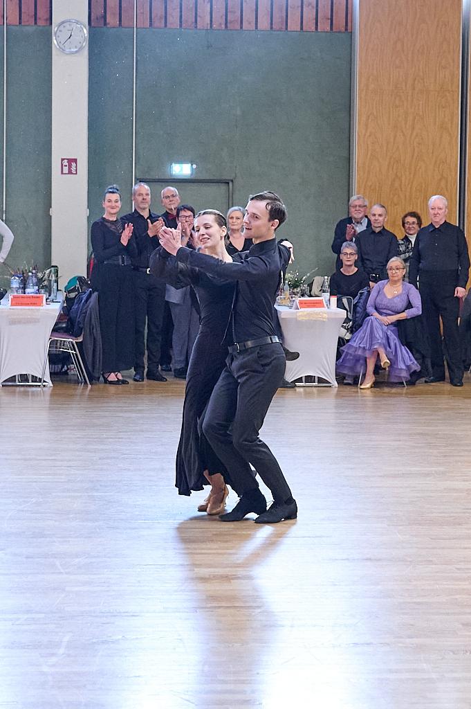 Tanzpaar in eleganter Kleidung tanzt auf einem Parkettboden, umgeben von applaudierendem Publikum.