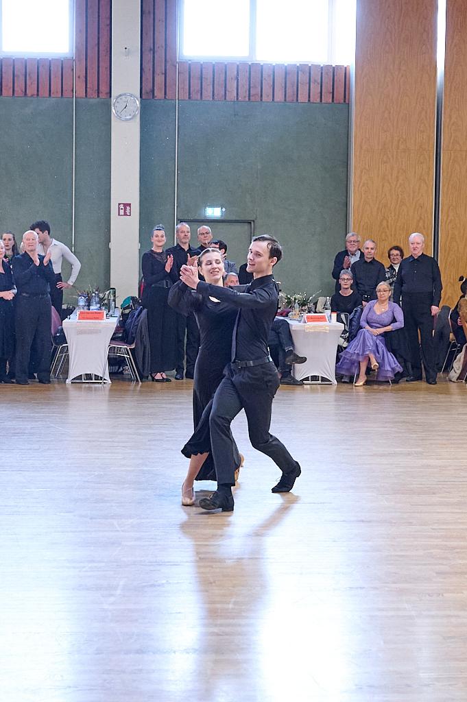 Tanzpaar in eleganter Kleidung tanzt auf einem Parkettboden vor applaudierendem Publikum in einem Saal.