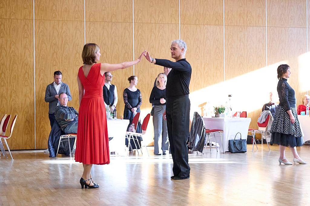 Tanzpaar in eleganter Kleidung f&uuml;hrt einen Schritt auf einer Tanzfl&auml;che, Zuschauer beobachten im Hintergrund.