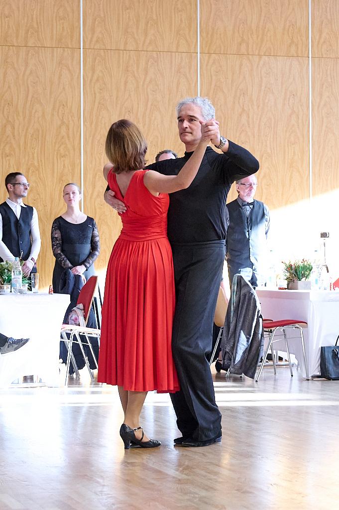 Tanzendes Paar in festlicher Kleidung, Frau im roten Kleid und Mann in schwarzem Oberteil, vor einer tanzenden Menge.