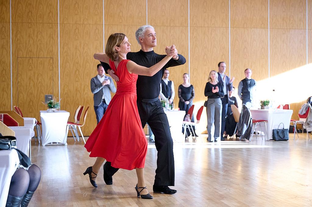 Tanzendes Paar in eleganter Kleidung, umgeben von Zuschauern in einem hellen Saal mit Holzboden.