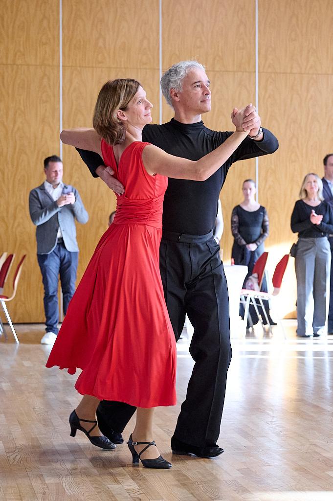 Tanzendes Paar in eleganter Kleidung, sie in rotem Kleid, er in schwarzem Oberteil, auf einem Parkettboden.