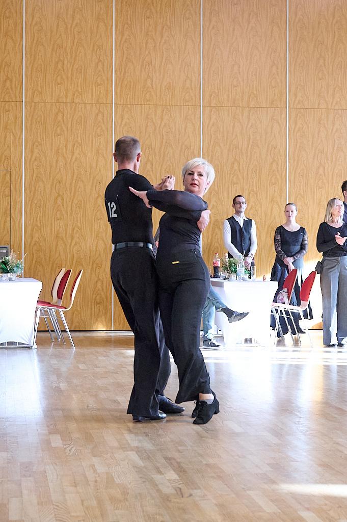 Tanzpaar f&uuml;hrt einen Schritt auf einer Tanzfl&auml;che, Zuschauer im Hintergrund beobachten die Vorf&uuml;hrung.