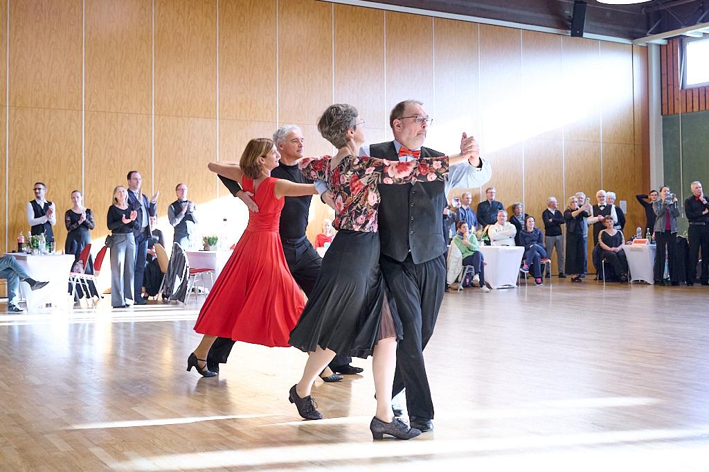 Tanzpaar in eleganter Kleidung f&uuml;hrt einen Tanz auf einer Holzbodenfl&auml;che, Zuschauer applaudieren im Hintergrund.