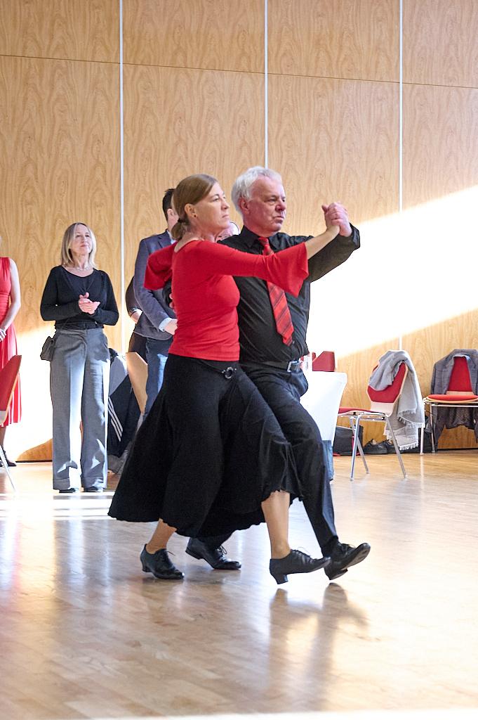 Tanzendes Paar in formeller Kleidung, umgeben von Zuschauern in einem hellen Saal mit Holzverkleidung.
