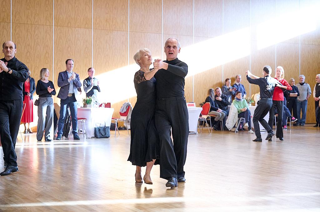 Tanzpaar in eleganter Kleidung tanzt in einer hellen Halle, Zuschauer applaudieren im Hintergrund.
