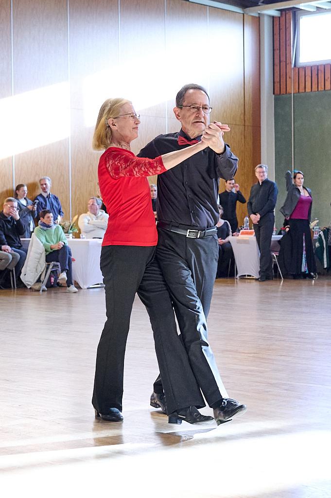 Tanzpaar in eleganter Kleidung tanzt miteinander auf einer hell erleuchteten Tanzfl&auml;che, Zuschauer im Hintergrund.