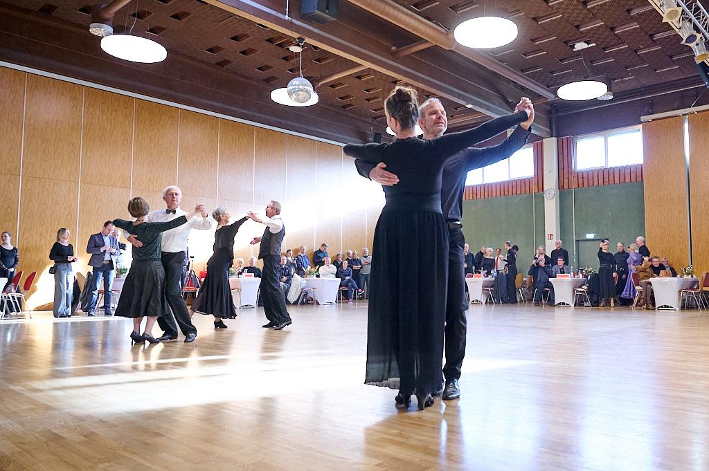 Tanzveranstaltung in einem Saal mit Paaren, die in eleganter Kleidung auf Holzfu&szlig;boden tanzen. Publikum schaut zu.