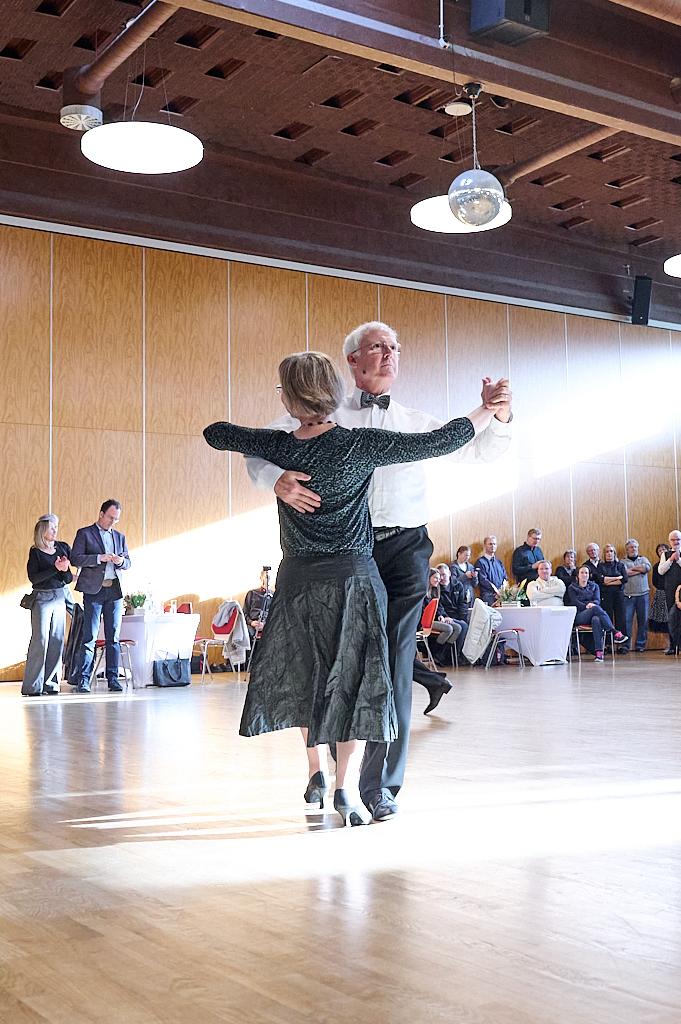 Tanzendes Paar in eleganter Kleidung auf einer Holzfl&auml;che, umgeben von Zuschauern bei einer festlichen Veranstaltung.
