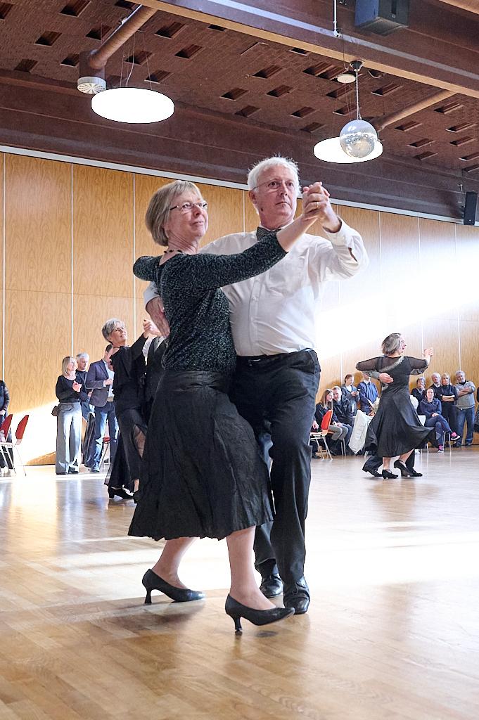 Tanzendes Paar in eleganter Kleidung auf einer Tanzfl&auml;che, umgeben von Zuschauern in einem hellen Raum.